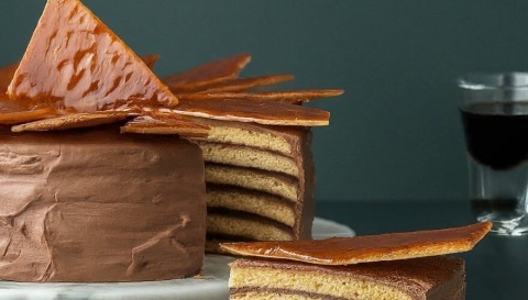 Doboš torta, isečeno parče torte i čaša sa pićem u pozadini.