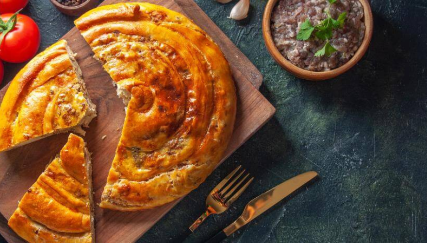 Burek sa mesom serviran na drvenoj dasci sa priborom za jelo pored.